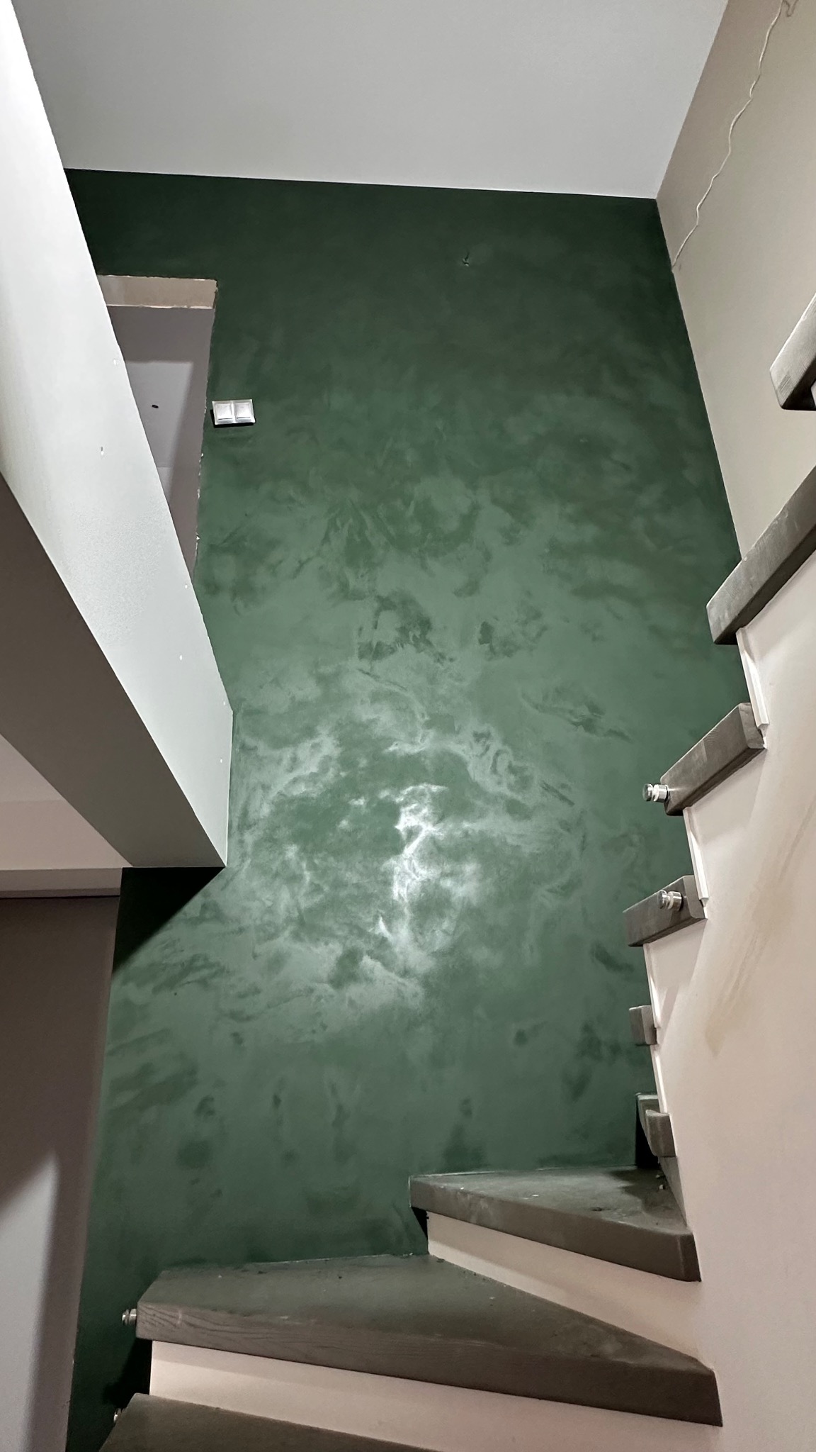 Ściana w odcieniach zieleni z efektem dekoracyjnym, widoczna klatka schodowa. Unikalna faktura farby tworzy subtelny, marmurkowy wzór. Oświetlenie podkreśla głębię koloru.