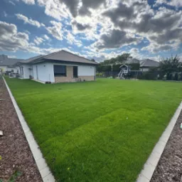 Zadbany trawnik przy nowoczesnym domu z placem zabaw w tle, obrzeża wykończone kostką i korą. Błękitne niebo z chmurami dodaje świeżości.