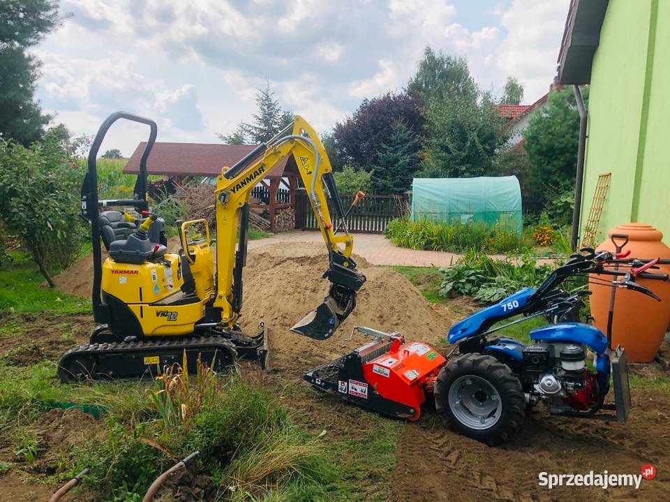Żółty mini-koparka Yanmar i glebogryzarka separacyjna w trakcie prac ziemnych we Wrocławiu. W tle widoczny ogród z tunelem foliowym i drewnianą altaną.