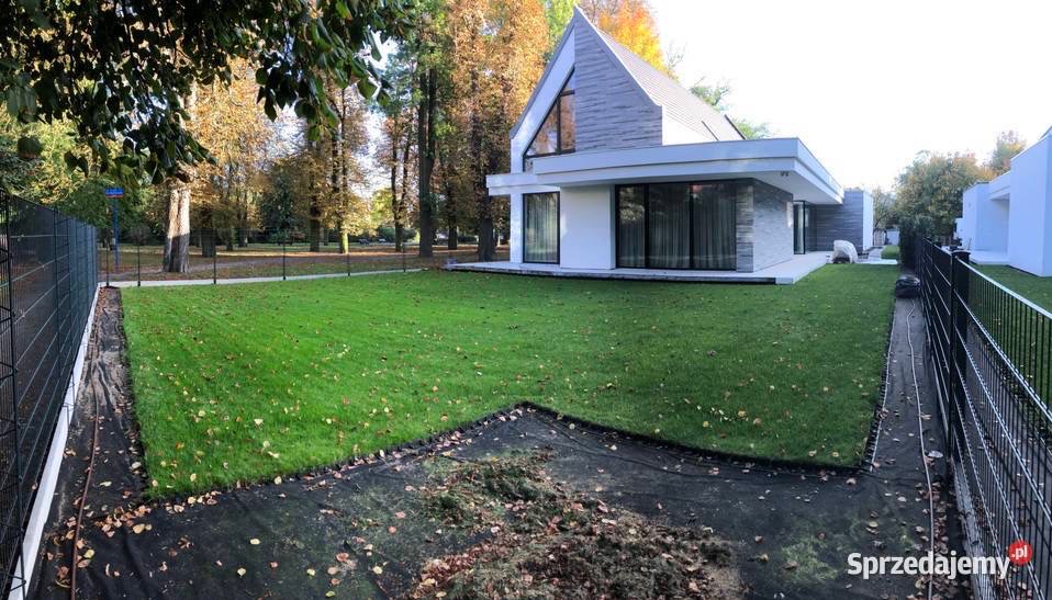 Nowoczesny dom z zadbanym trawnikiem; widoczne prace przygotowawcze na obrzeżach, siatka zabezpieczająca, jesienne drzewa w tle.