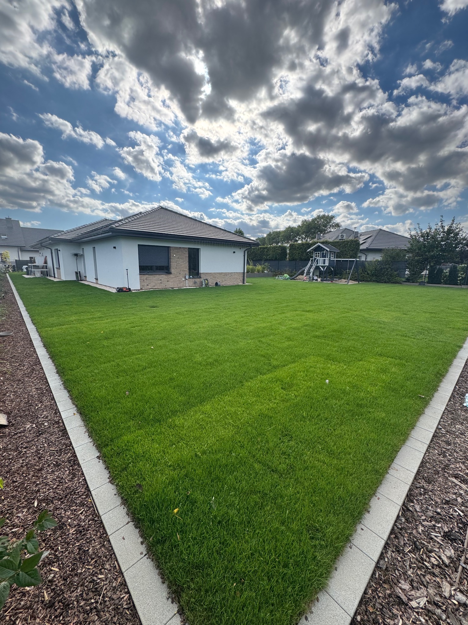 Zadbany trawnik przy nowoczesnym domu z placem zabaw w tle, obrzeża wykończone kostką i korą. Błękitne niebo z chmurami dodaje świeżości.