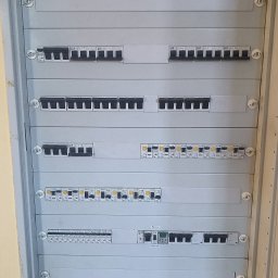 JAGO Grzegorz Brzoza - Rozdzielnica elektryczna z bezpiecznikami i wyłącznikami, widok z bliska. Szara obudowa, widoczne oznaczenia i przewody. Element instalacji elektrycznej.