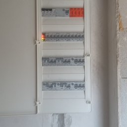 JAGO Grzegorz Brzoza - Otwarta skrzynka bezpiecznikowa z włączonymi bezpiecznikami, zamontowana na surowej ścianie we Wrocławiu. Widoczne kable i elementy instalacji elektrycznej.