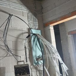 JAGO Grzegorz Brzoza - Surowy stan instalacji elektrycznej w budynku z białymi kablami ułożonymi w rurkach i czarnymi kablami zwisającymi luźno, widoczne puszki elektryczne i ściany z betonu komórkowego.