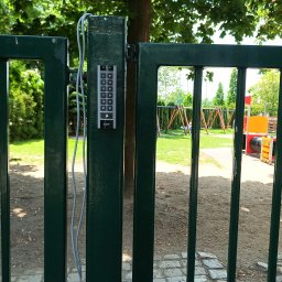 JAGO Grzegorz Brzoza - Zielona metalowa furtka z zamontowanym panelem numerycznym dostępu, widok na plac zabaw w tle, słoneczny dzień.