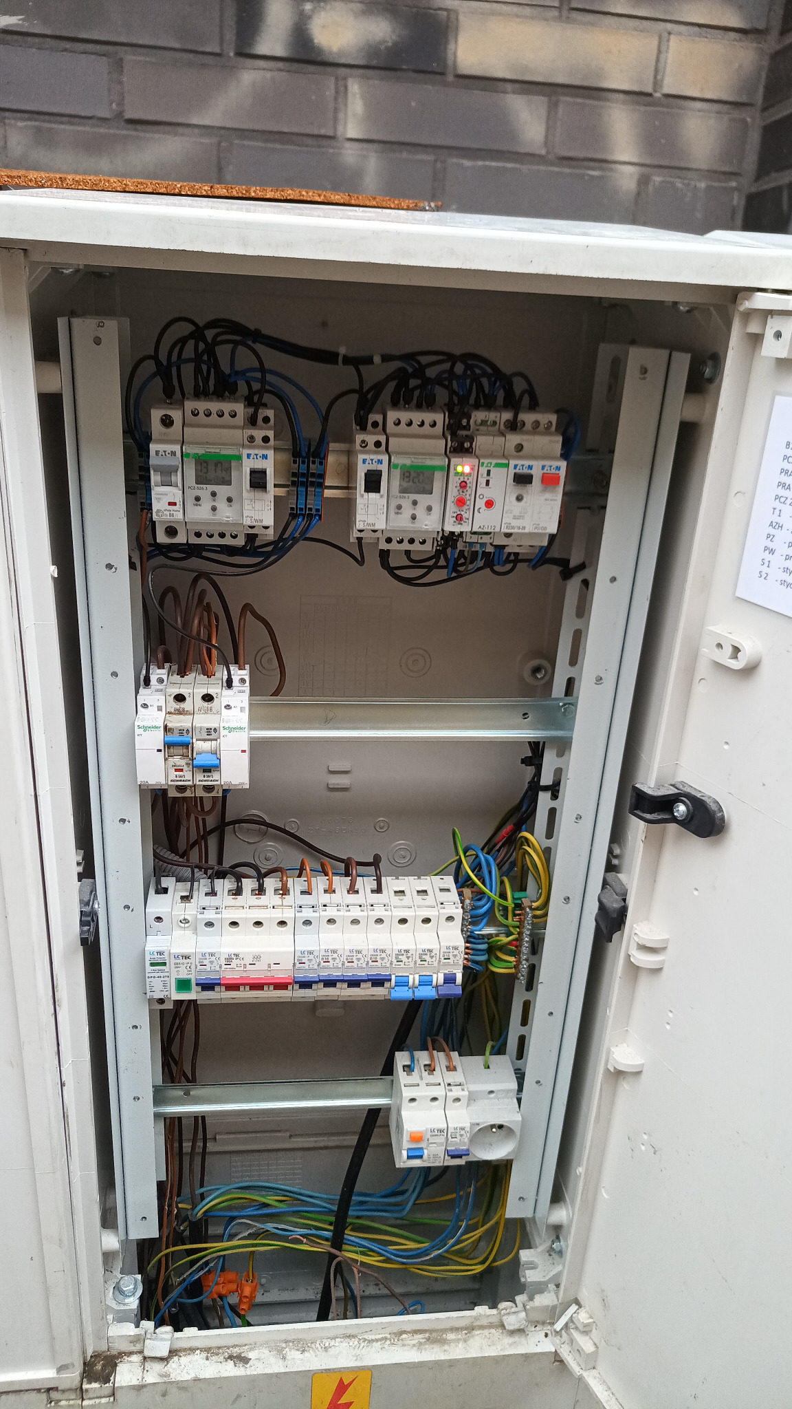 Otwarta skrzynka elektryczna z zainstalowanymi bezpiecznikami, przewodami i elementami sterującymi. Widoczne komponenty Eaton i Schneider Electric.