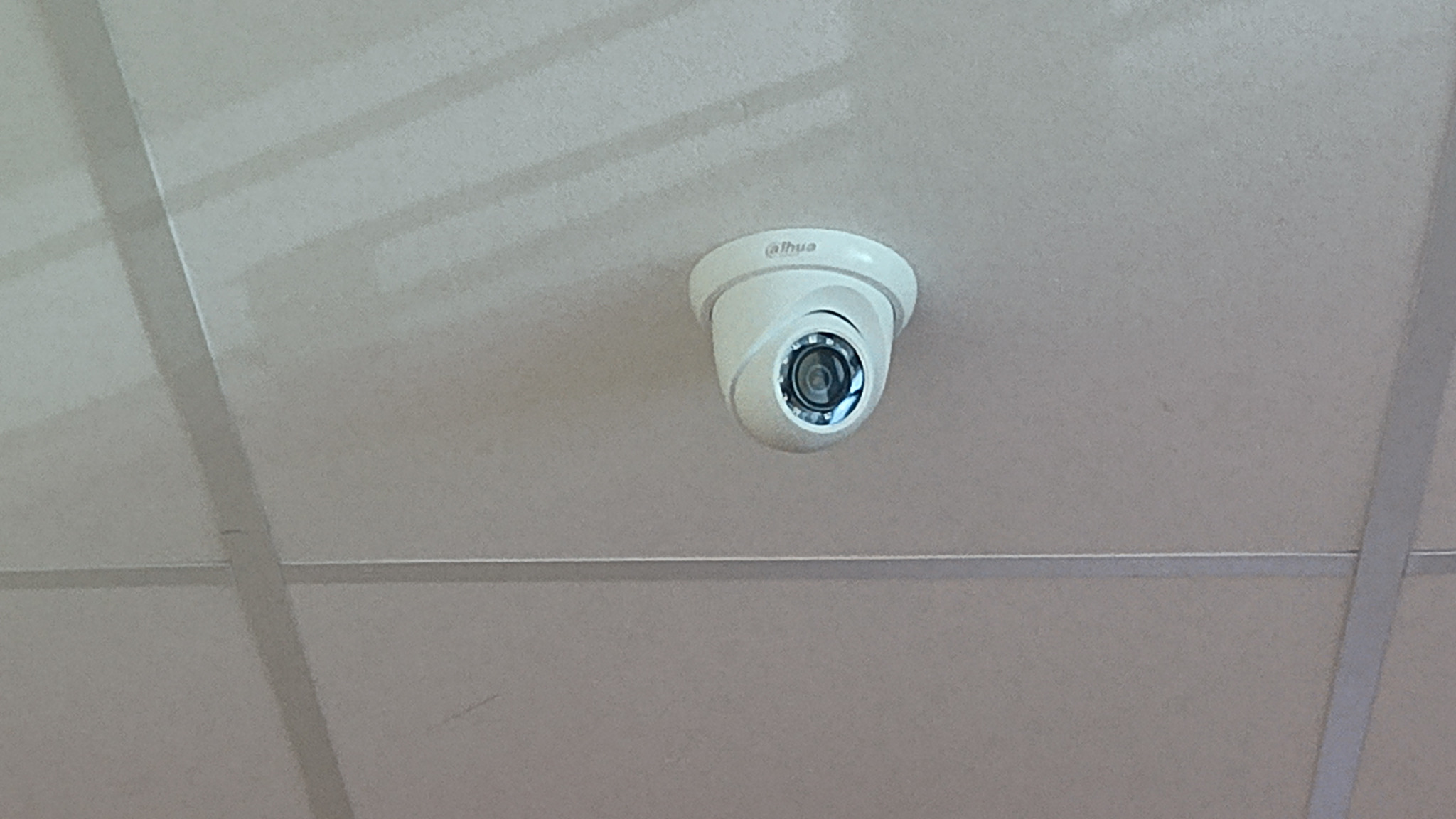 Kopułkowa kamera monitoringu Dahua zamontowana na suficie kasetonowym, skierowana obiektywem w dół. Widoczne delikatne cienie i nierówności sufitu.