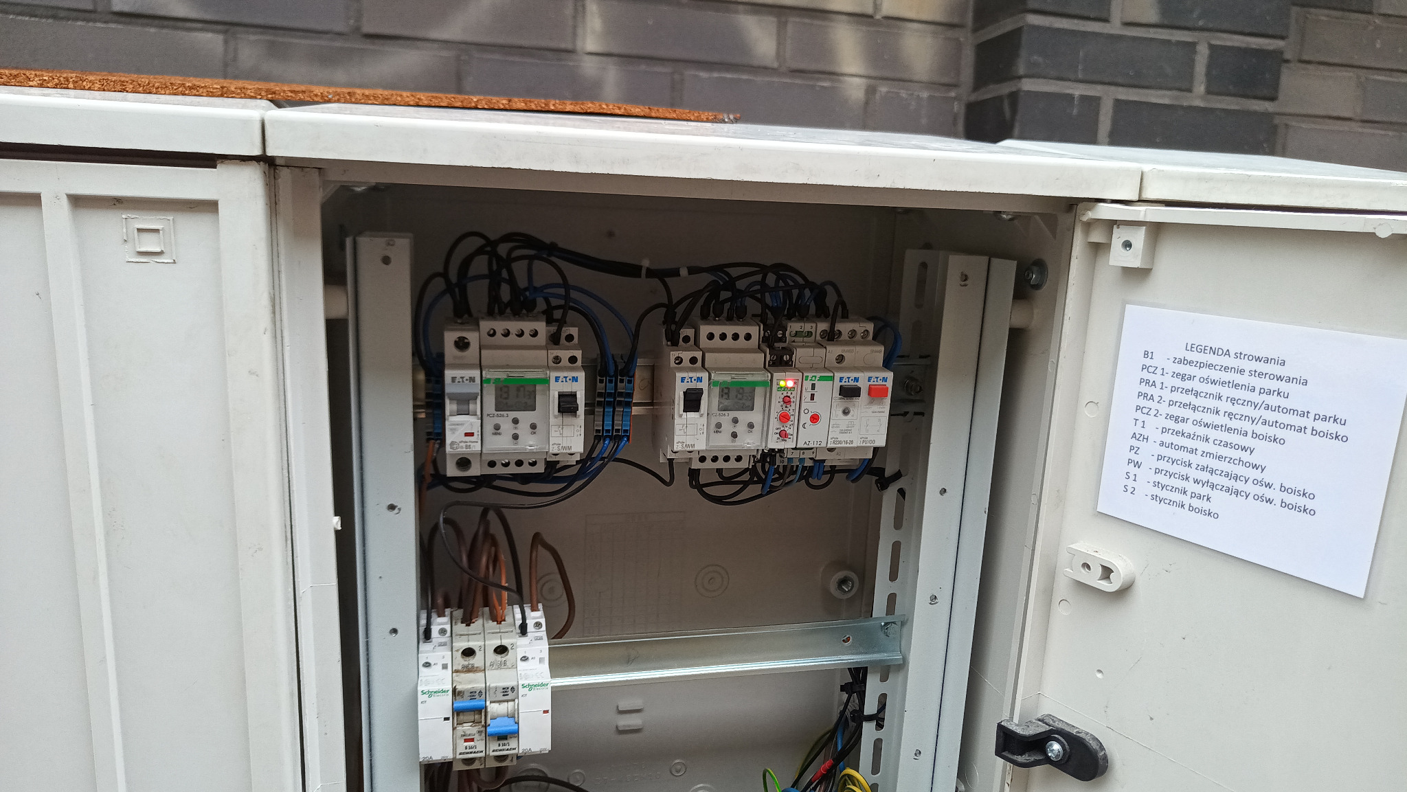 Otwarta skrzynka elektryczna z zabezpieczeniami, zegarami sterującymi oświetleniem parku i boiska. Widoczne elementy sterowania i legenda opisująca funkcje.