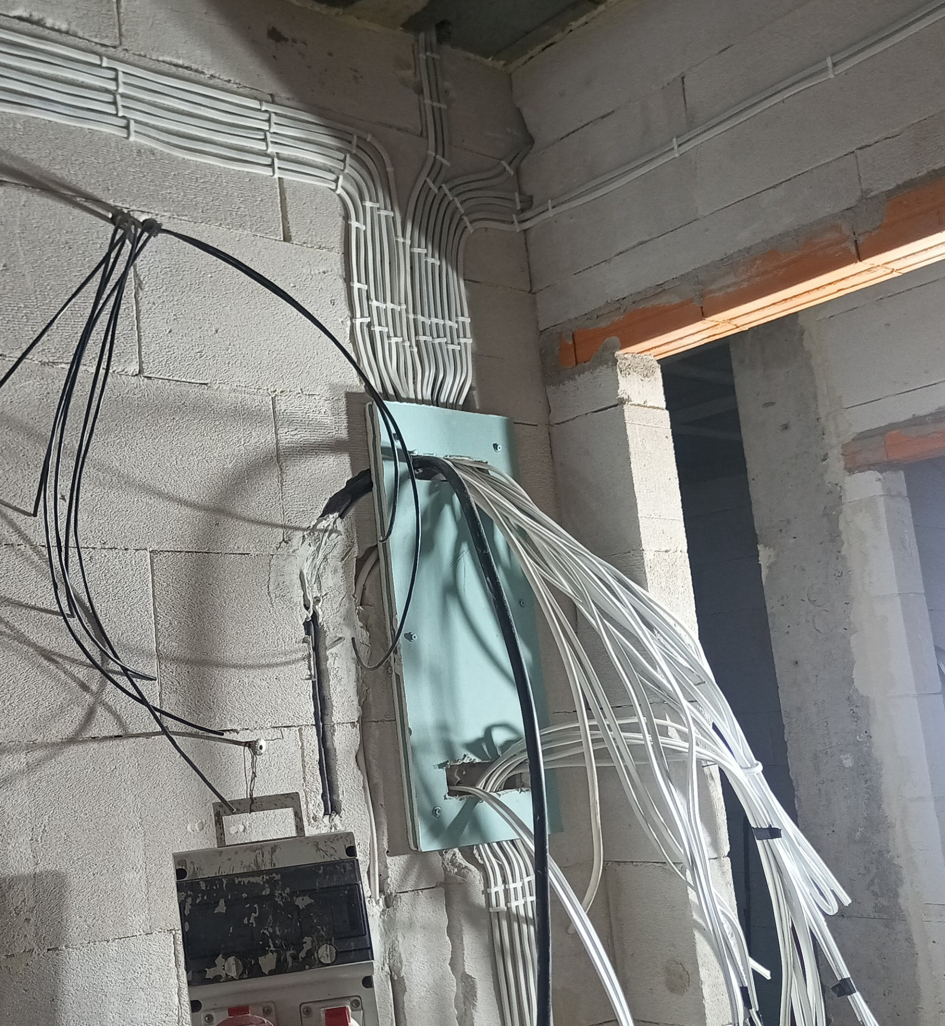 Surowy stan instalacji elektrycznej w budynku z białymi kablami ułożonymi w rurkach i czarnymi kablami zwisającymi luźno, widoczne puszki elektryczne i ściany z betonu komórkowego.