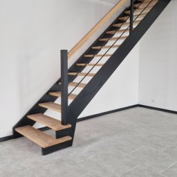 Zakład Stolarski Katarzyna Czyż - Nowoczesne schody z jasnego drewna i czarnej stali w minimalistycznym wnętrzu. Widok z dołu ukazuje konstrukcję i subtelne oświetlenie.