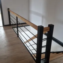 Nowoczesna balustrada schodowa z drewnianym poręczą i czarnymi, metalowymi prętami, widok z góry na drewniane schody i podłogę.