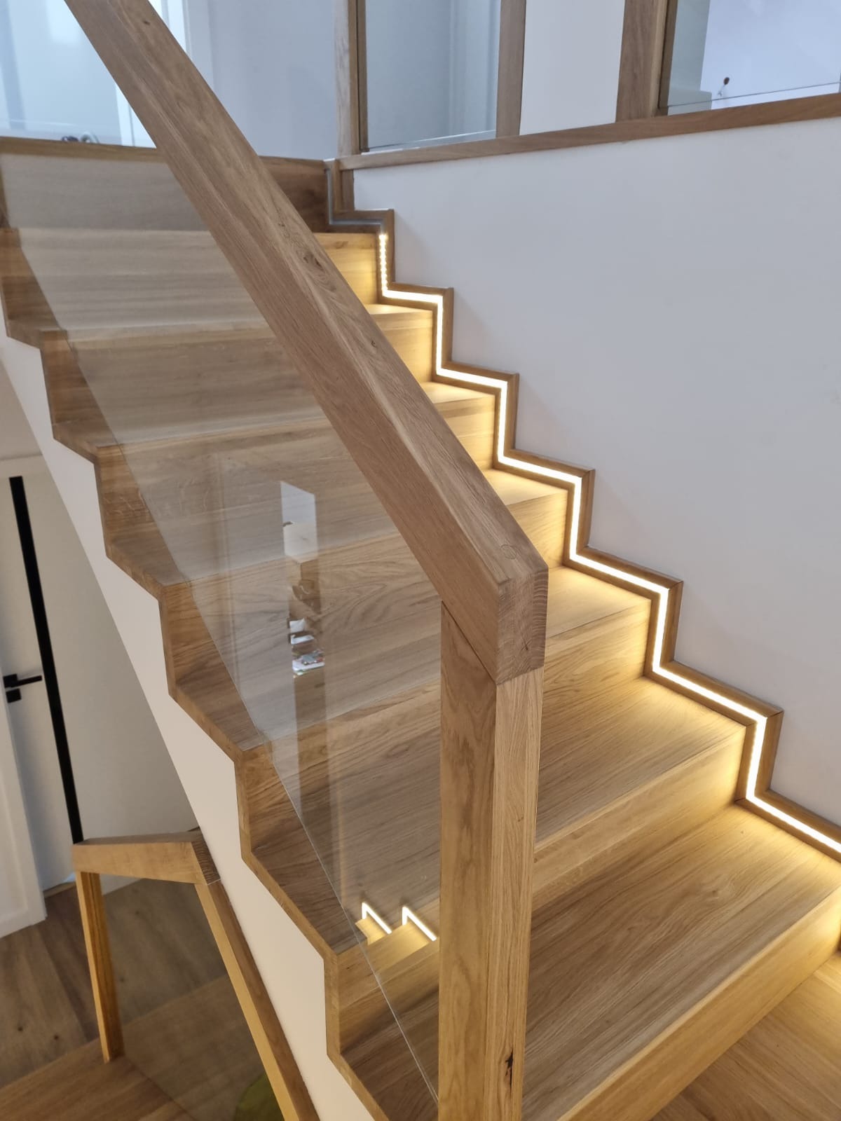 Nowoczesne schody z drewna z oświetleniem LED wzdłuż stopni i szklaną balustradą. Jasne drewno, minimalistyczny design, subtelne oświetlenie, schody w domu.