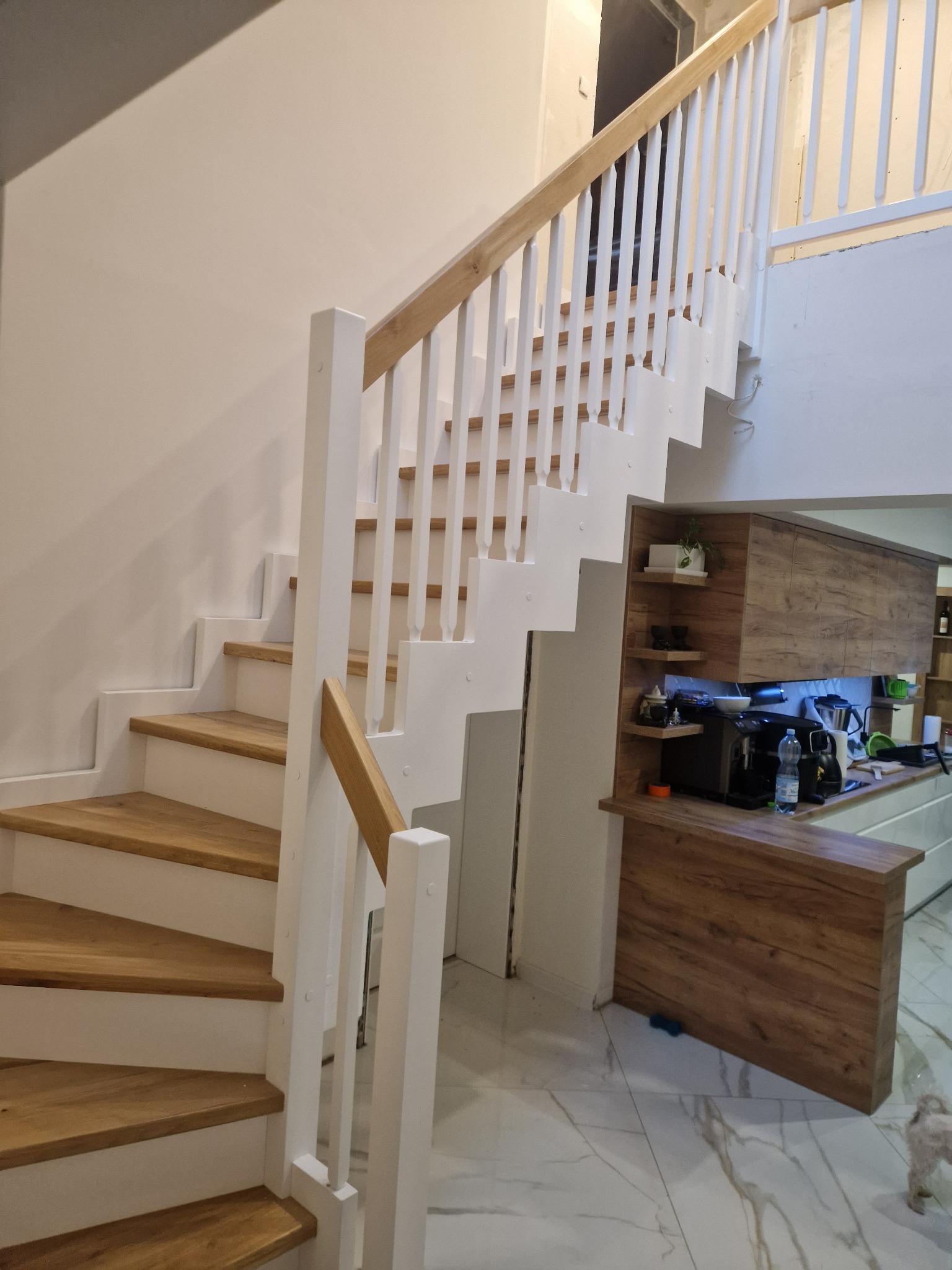 Nowoczesne schody z drewnianymi stopniami i białą balustradą w jasnym wnętrzu, widok z dołu, fragment zabudowy kuchennej w tle.