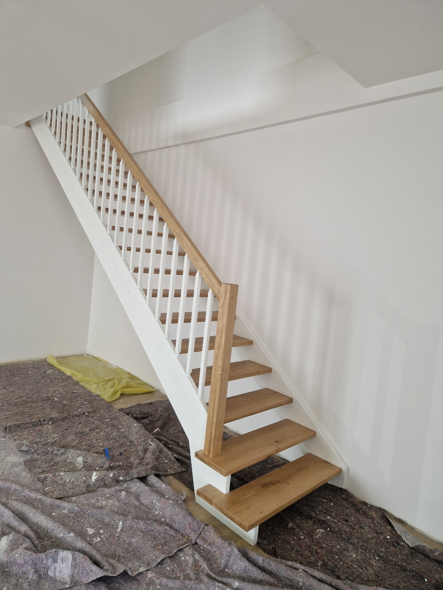 Wnętrze z drewnianymi schodami o jasnych stopniach i białej balustradzie. Na podłodze zabezpieczenie malarskie. Minimalistyczny design w nowym budownictwie.