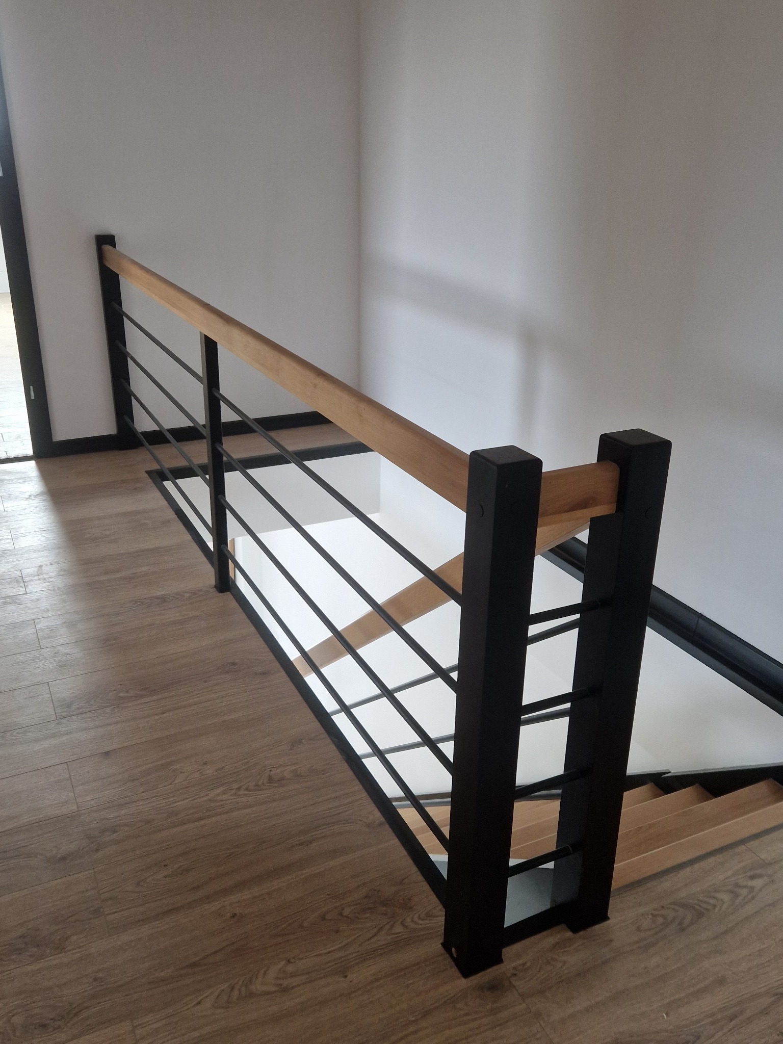 Nowoczesna balustrada schodowa z drewnianym poręczą i czarnymi, metalowymi prętami, widok z góry na drewniane schody i podłogę.