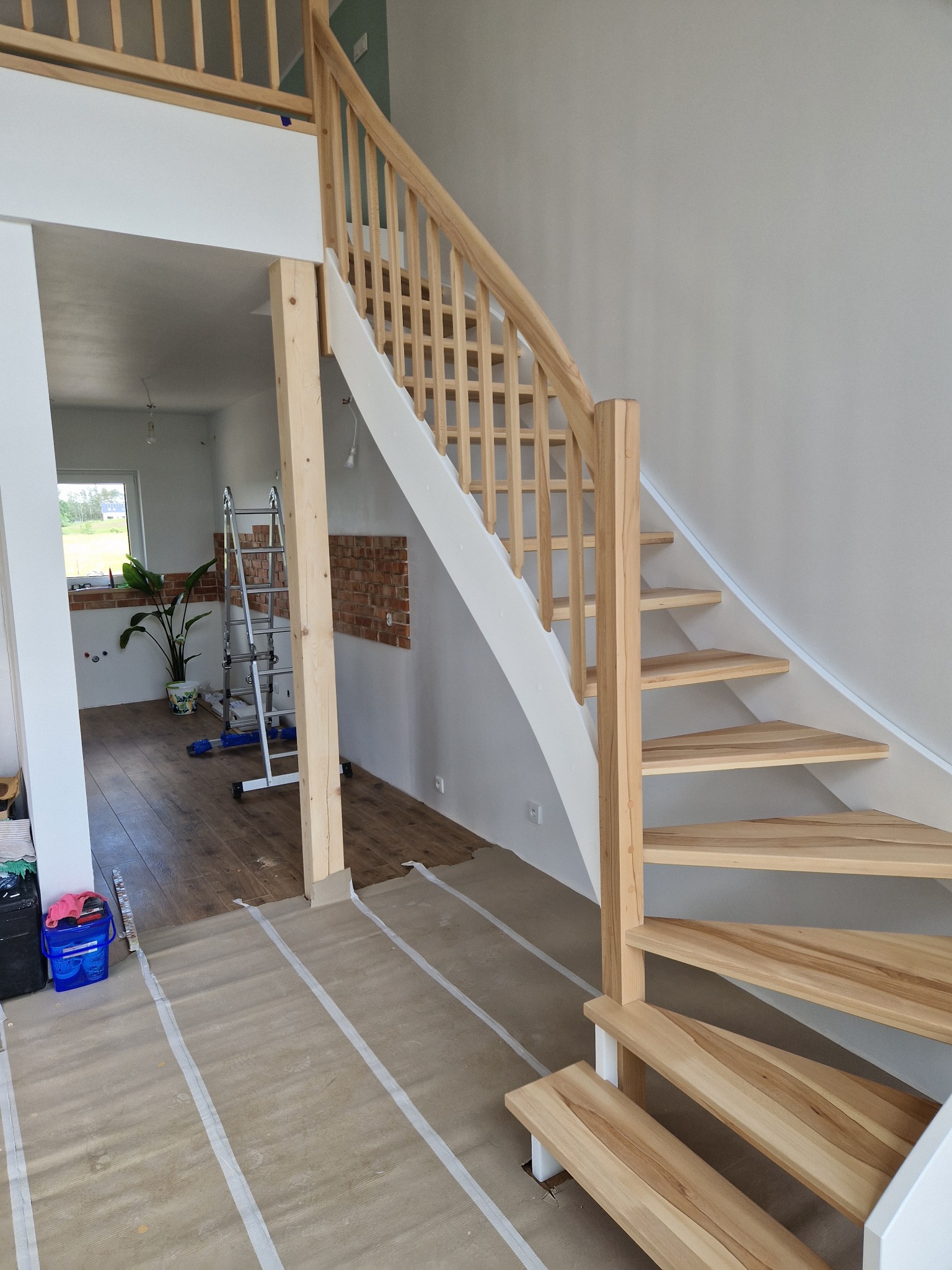 Jasne, drewniane schody z białą balustradą w surowym wnętrzu. Widoczna drabina i przygotowane podłoże, co sugeruje etap montażu lub wykończenia.