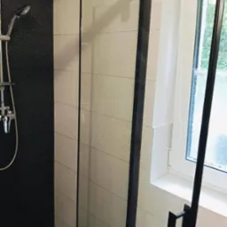 Nowoczesna kabina prysznicowa z czarnymi ramami i deszczownicą, wykończona płytkami w odcieniach beżu i czerni, z oknem w tle. Detal łazienki po remoncie.