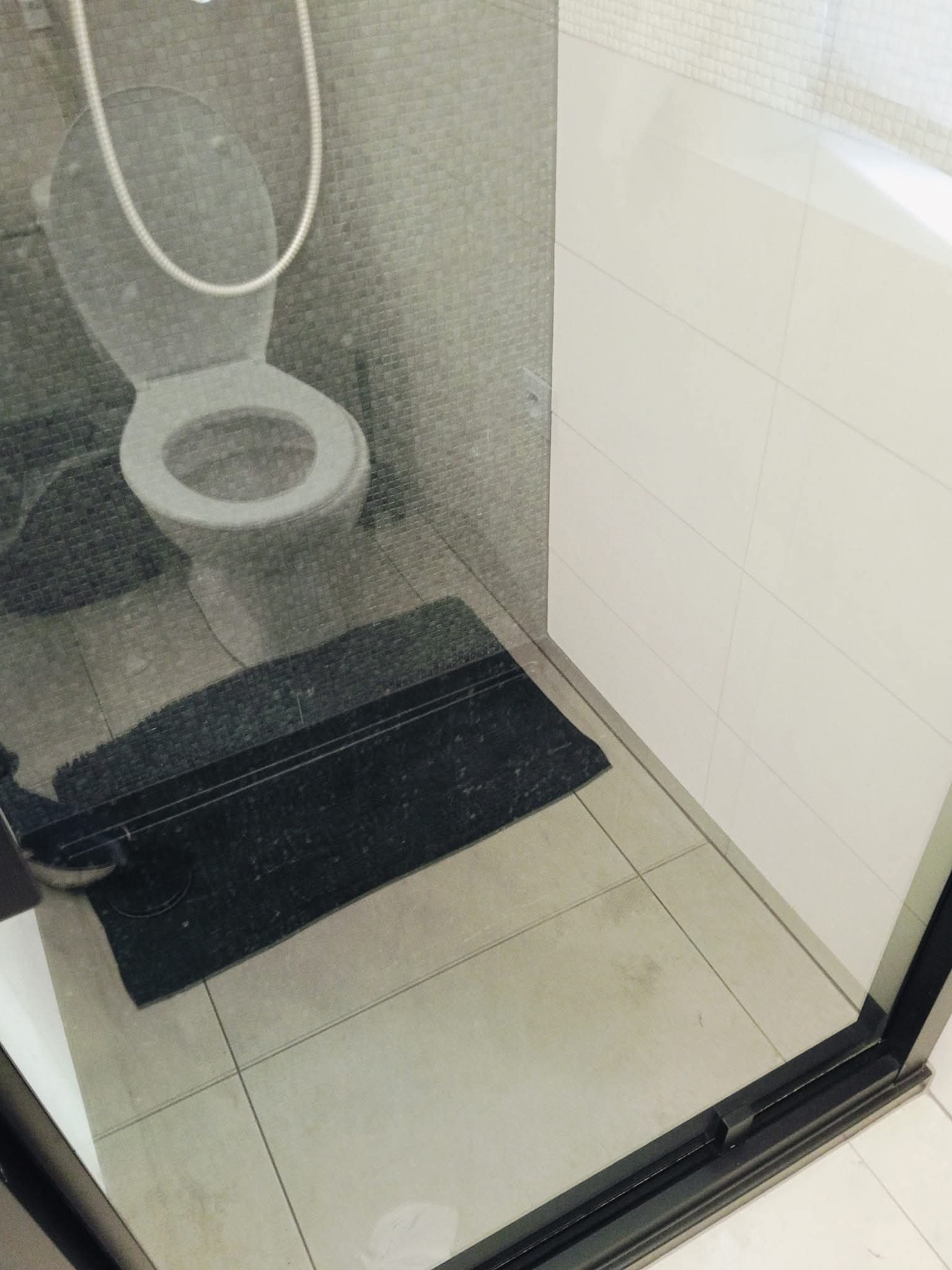 Wnętrze łazienki z toaletą i szarym dywanikiem, widoczne przez szybę kabiny prysznicowej. Minimalistyczny design, płytki w odcieniach szarości i bieli.