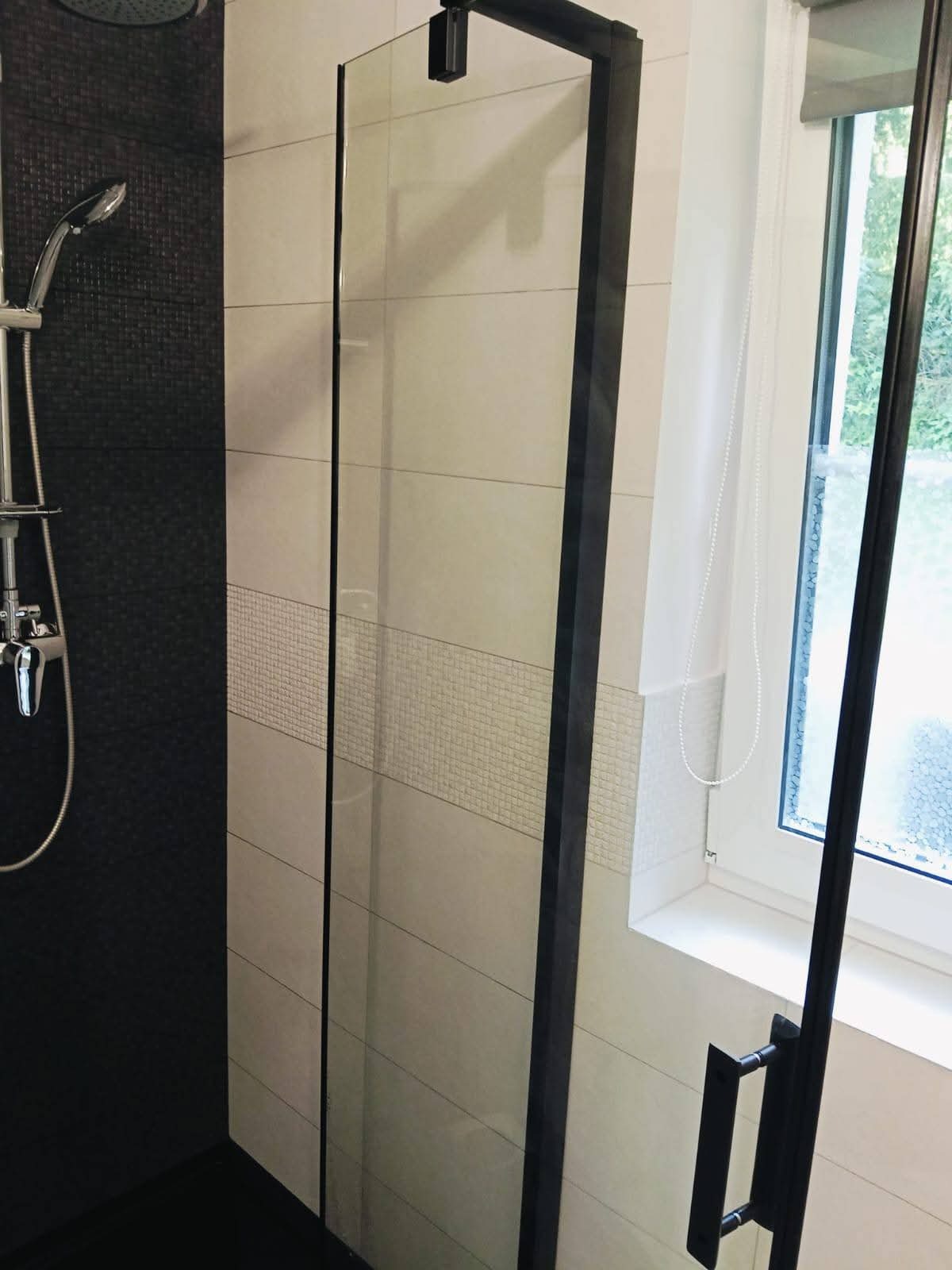 Nowoczesna kabina prysznicowa z czarnymi ramami i deszczownicą, wykończona płytkami w odcieniach beżu i czerni, z oknem w tle. Detal łazienki po remoncie.