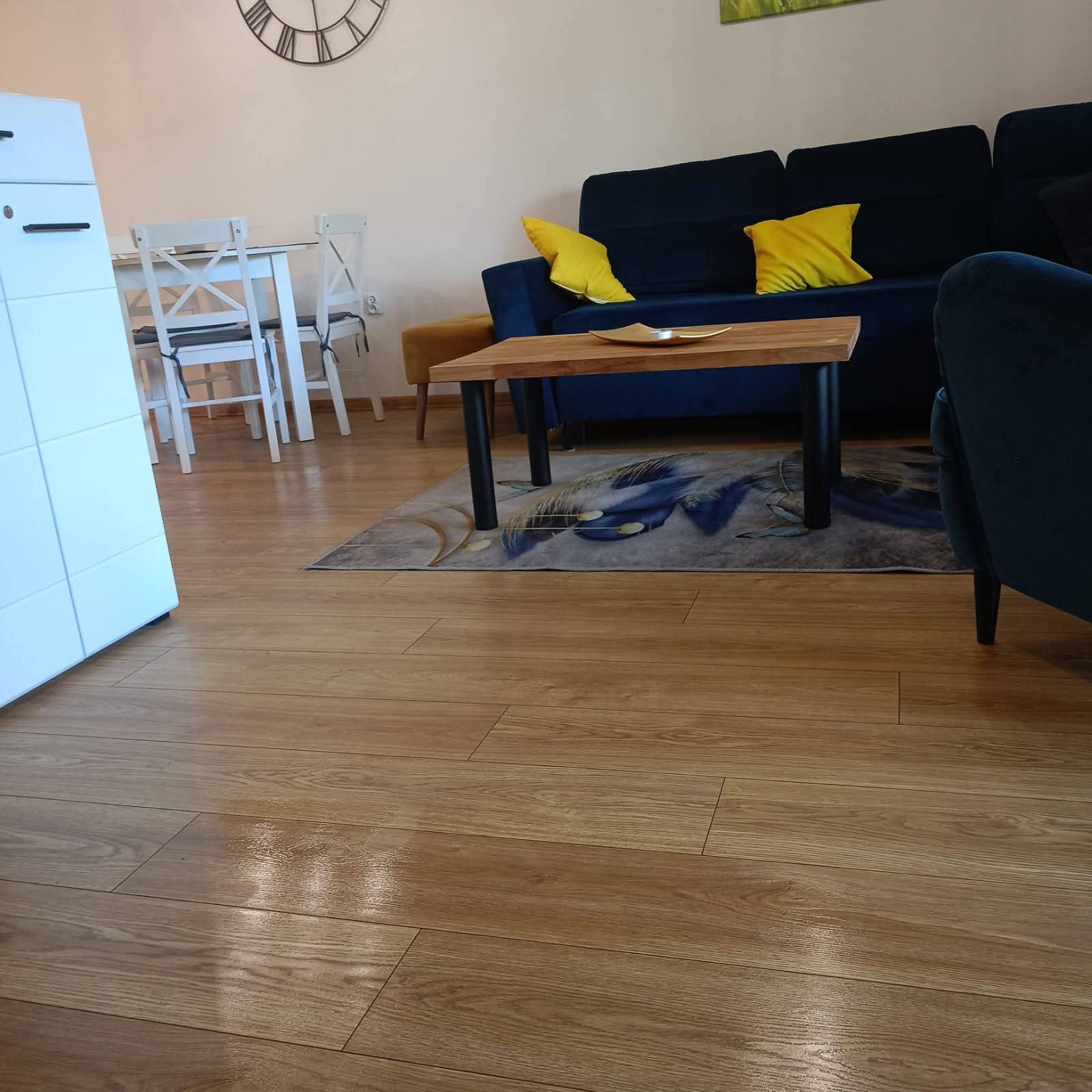 Salon z drewnianą podłogą, niebieską sofą z żółtymi poduszkami, stolikiem kawowym i białymi krzesłami. W tle zegar na ścianie. Widok na czystą i zadbaną przestrzeń.