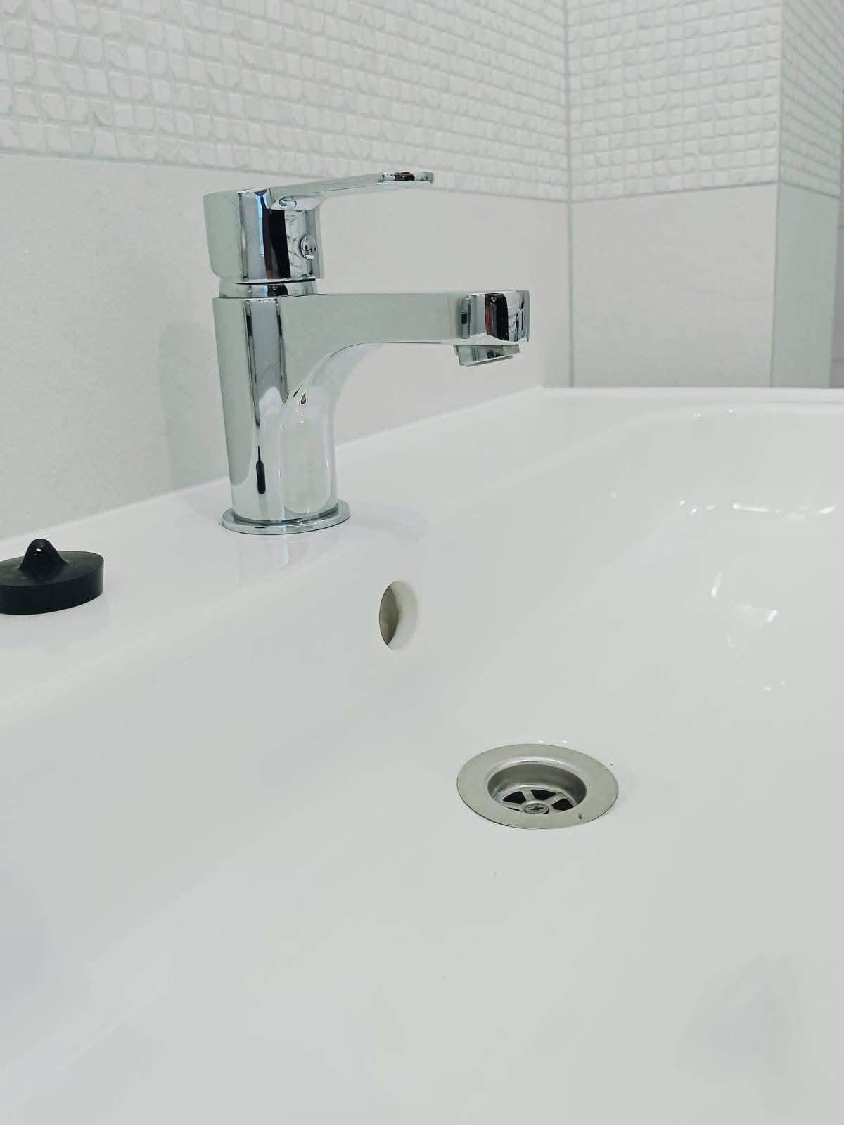 Biała umywalka z chromowaną baterią i korkiem. Świeżo po montażu, na ścianie białe płytki z delikatną fakturą. Widok z góry, ostre światło.