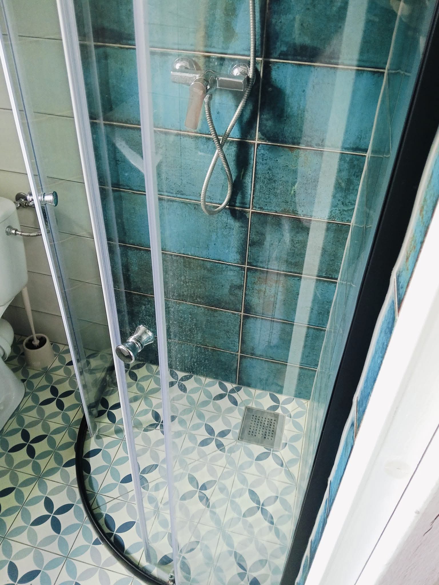 Nowoczesna kabina prysznicowa z turkusowymi płytkami i wzorzystą podłogą. Widoczny chromowany zestaw prysznicowy i odpływ liniowy. Elementy toalety w tle.