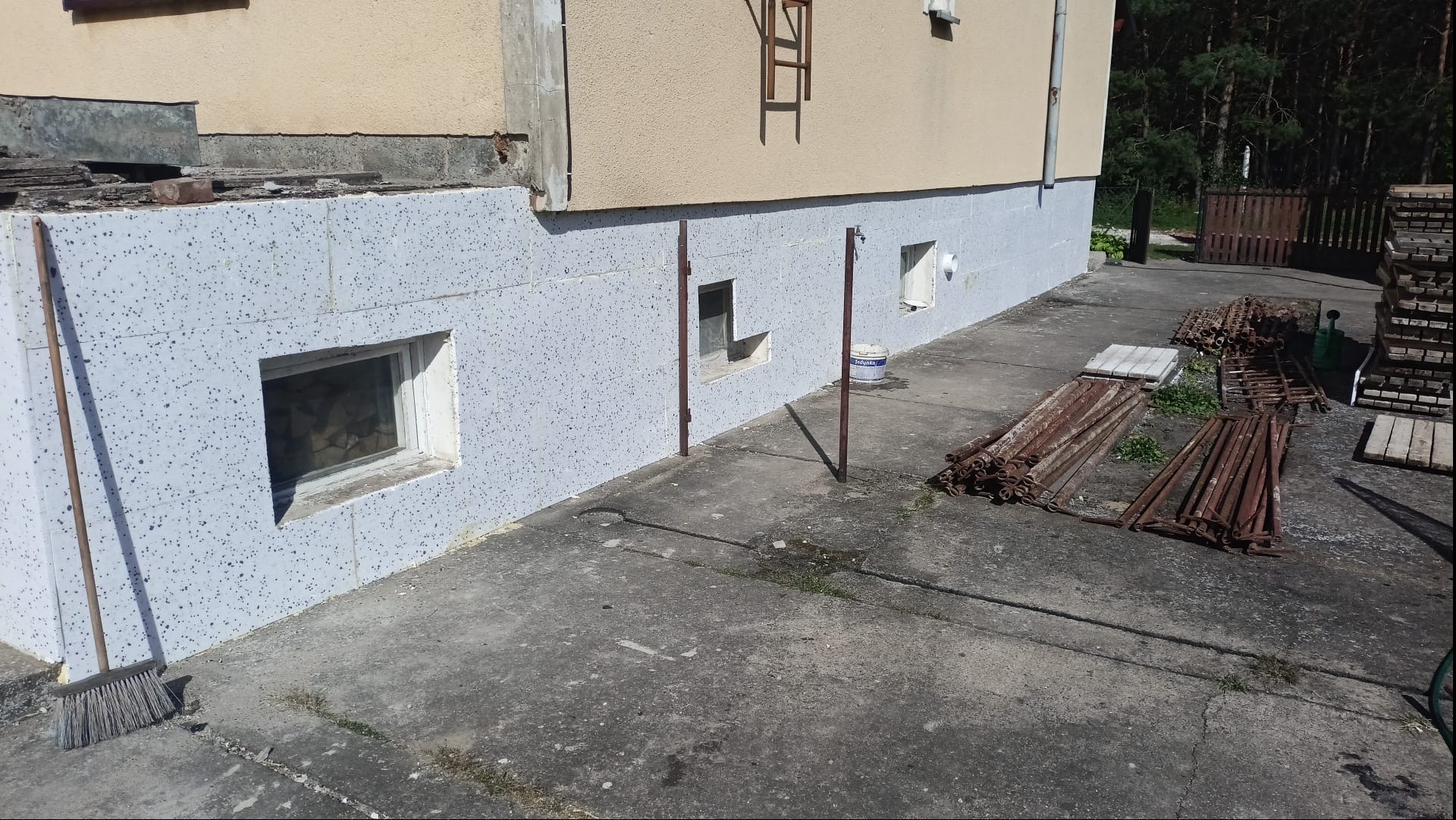 Fragment budynku po ociepleniu elewacji z widocznym styropianem, oknami piwnicznymi i stertą rusztowań na zewnątrz. Widoczny fragment elewacji w kolorze kremowym.