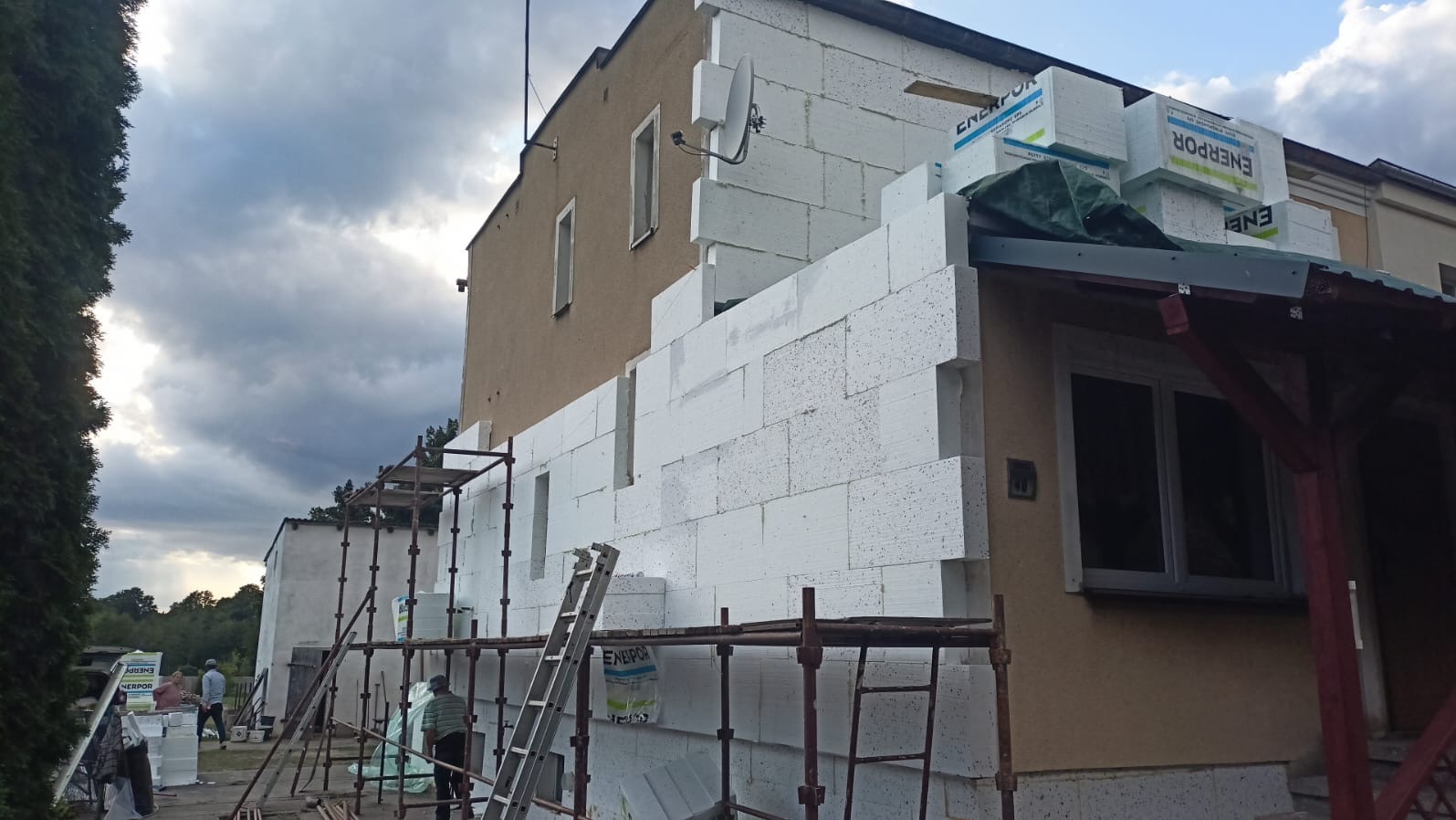 Budynek w trakcie ocieplania styropianem, widoczne rusztowania i ekipa. Częściowo ocieplona elewacja budynku z zamontowaną anteną satelitarną.