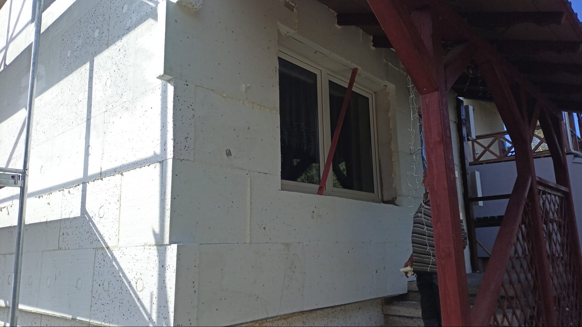 Biała elewacja budynku w trakcie ocieplania, widoczne płyty styropianowe i okno z czerwoną poziomicą. Fragment rusztowania po lewej stronie.