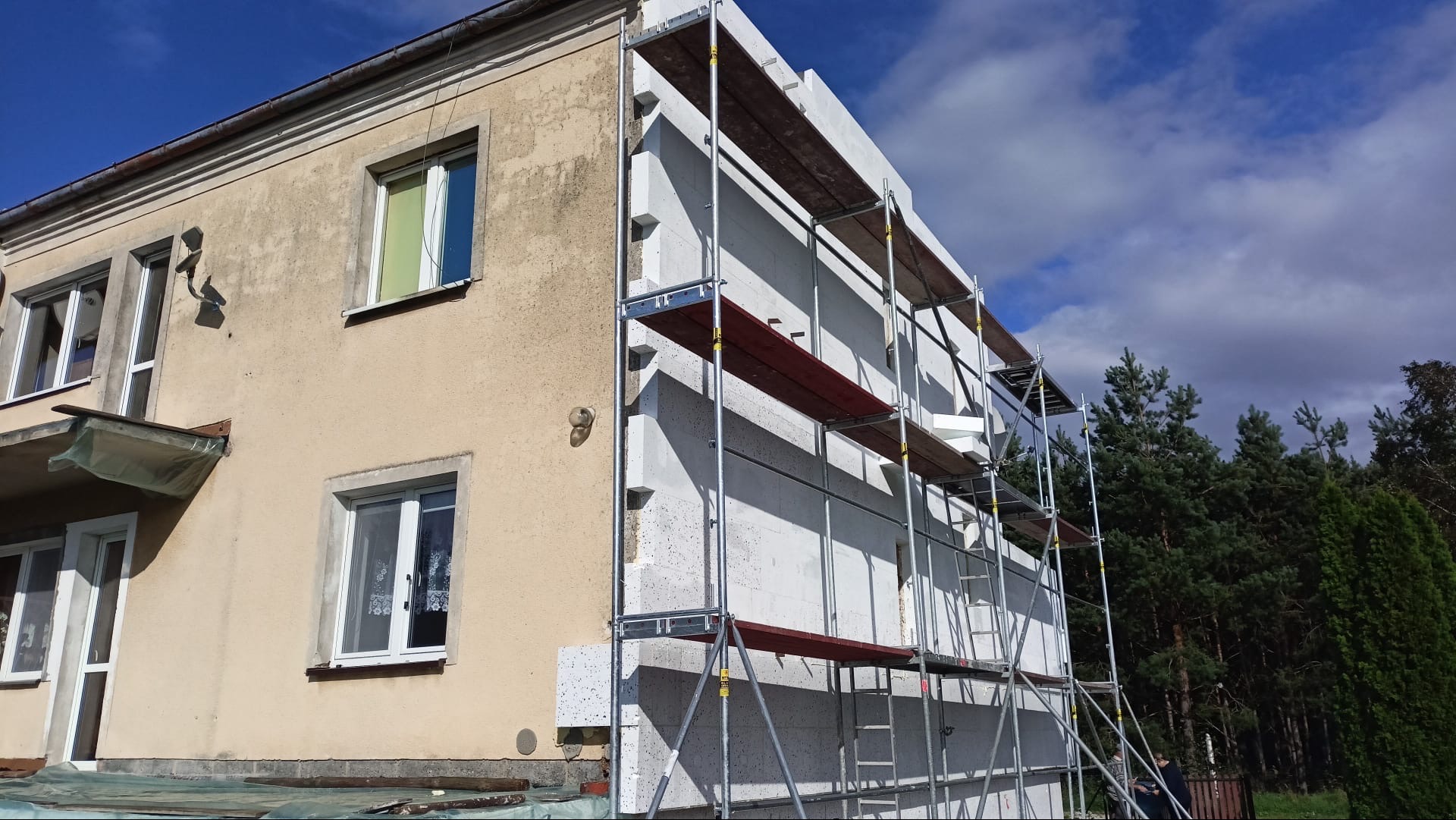 Dom w trakcie ocieplania: białe płyty izolacyjne na elewacji, rusztowania. Część budynku otynkowana, z oknami. W tle zieleń drzew i błękitne niebo.