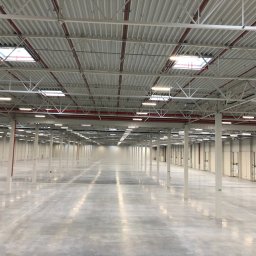 Stecom Lighting Sp. z o.o. - Wnętrze hali magazynowej z równomiernie rozmieszczonymi lampami sufitowymi, widoczne białe kolumny i szara posadzka. Oświetlenie równomierne, bez wyraźnych cieni.