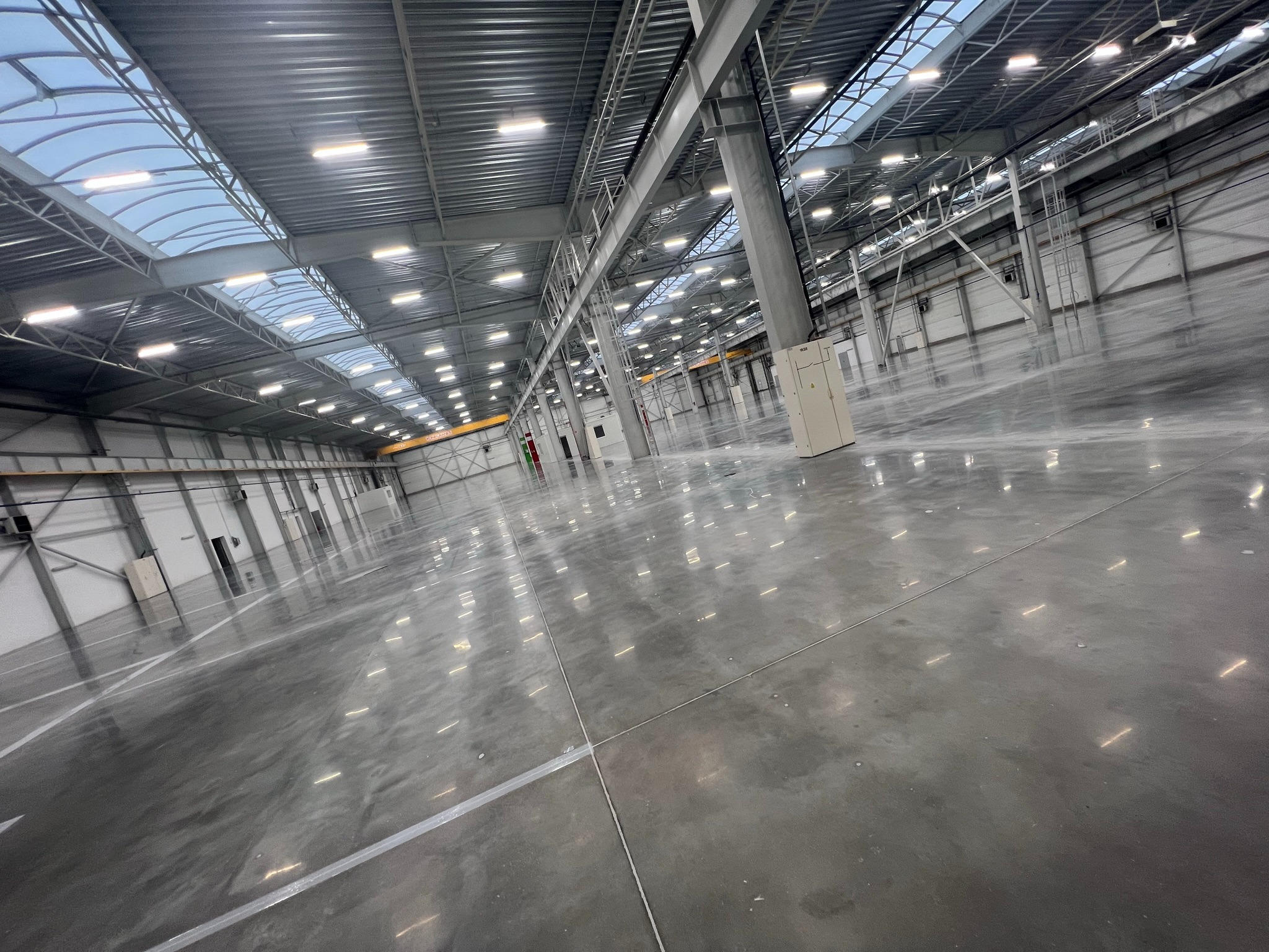 Wnętrze hali magazynowej z zamontowanym oświetleniem LED. Widoczne świetliki dachowe i betonowa posadzka z wyznaczonymi liniami. Perspektywa z niskiego kąta.