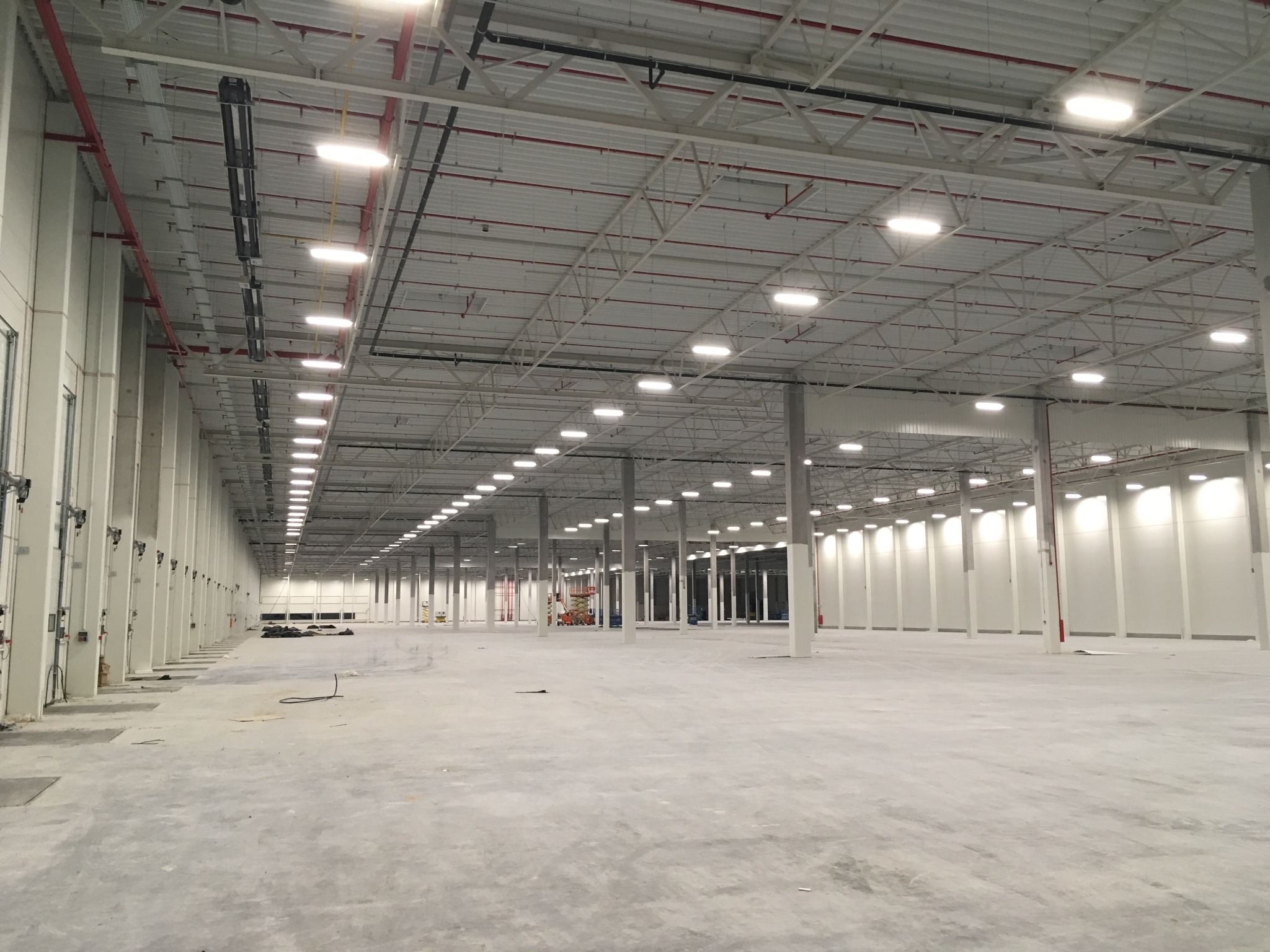 Wnętrze hali magazynowej z równomiernie rozmieszczonymi lampami sufitowymi, widoczne instalacje pod stropem i białe kolumny. Hala przygotowana do użytku.