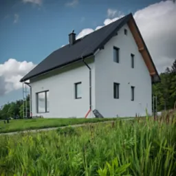 Nowoczesny dom jednorodzinny z czarnym dachem, jasną elewacją i czarnymi ramami okiennymi, otoczony zielenią. Widoczne rusztowania sugerują prace wykończeniowe.