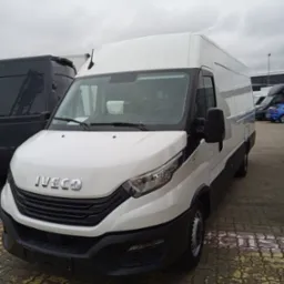 Biały bus Iveco z widoczną kartką za szybą, stojący na parkingu z kostki brukowej w pochmurny dzień, z innymi pojazdami w tle.