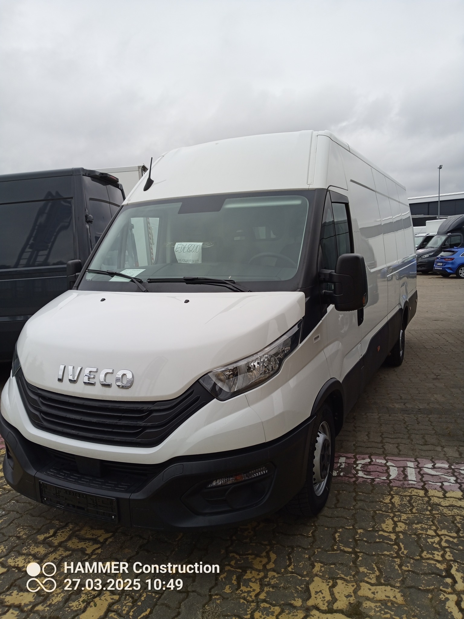 Biały bus Iveco z widoczną kartką za szybą, stojący na parkingu z kostki brukowej w pochmurny dzień, z innymi pojazdami w tle.