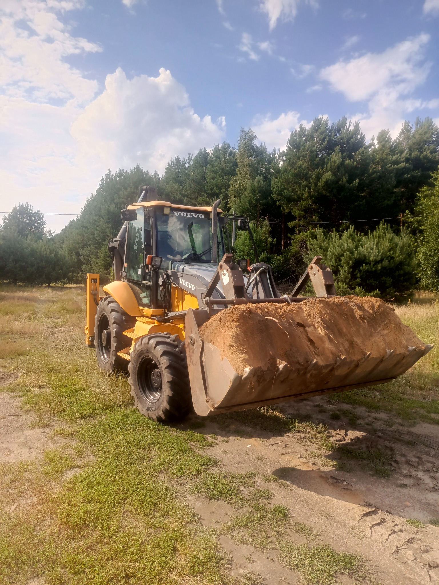 Żółto-szara koparko-ładowarka Volvo z pełną łyżką piasku na tle drzew i błękitnego nieba. Sprzęt budowlany w naturalnym otoczeniu.