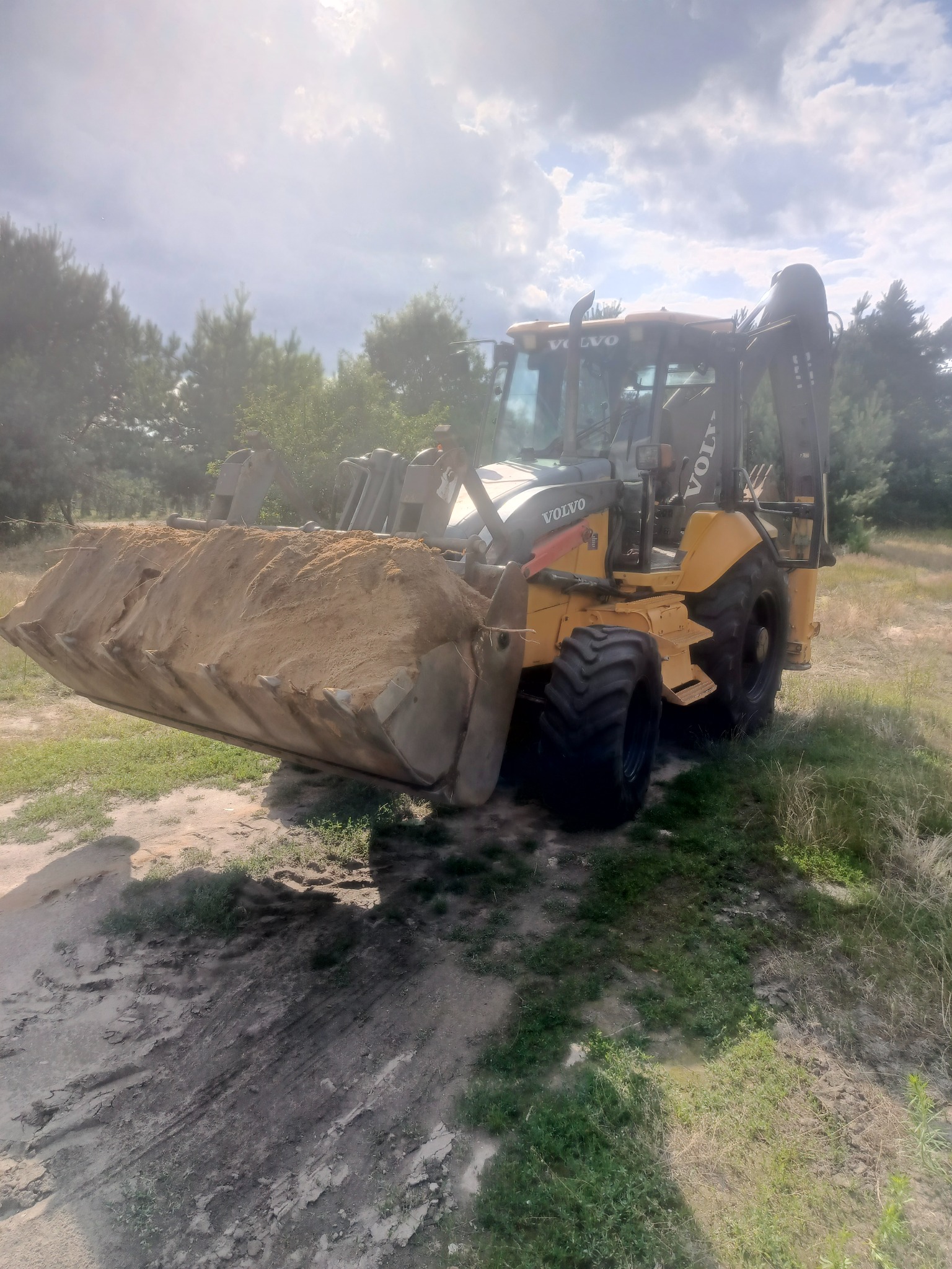 Żółta koparko-ładowarka Volvo z pełną łyżką piasku na tle drzew i pochmurnego nieba. Widoczne ślady opon na ziemi.