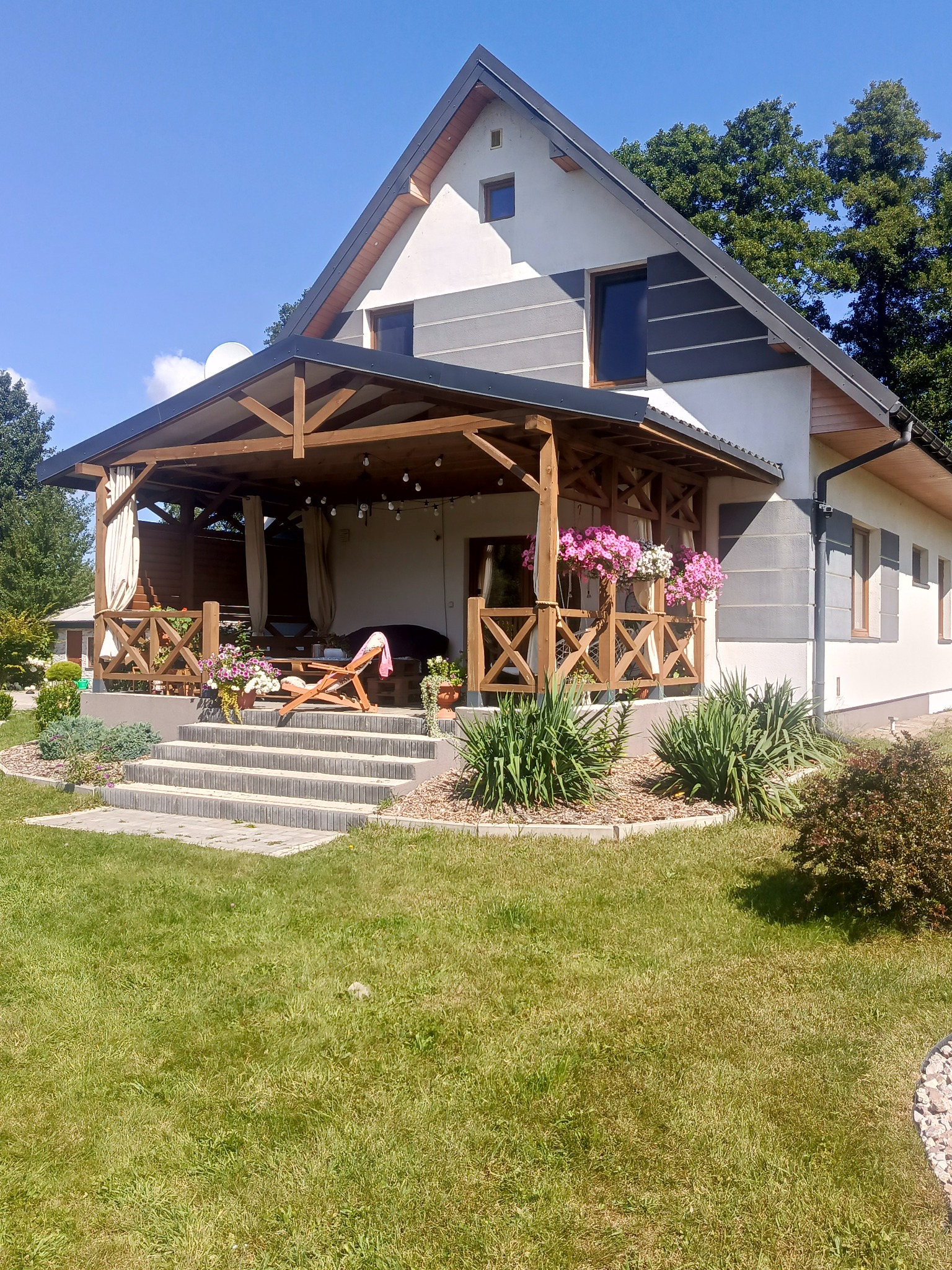 Nowoczesny dom z drewnianą werandą, schodami z kostki i zadbanym trawnikiem w słoneczny dzień. Kwiaty w donicach dodają uroku.