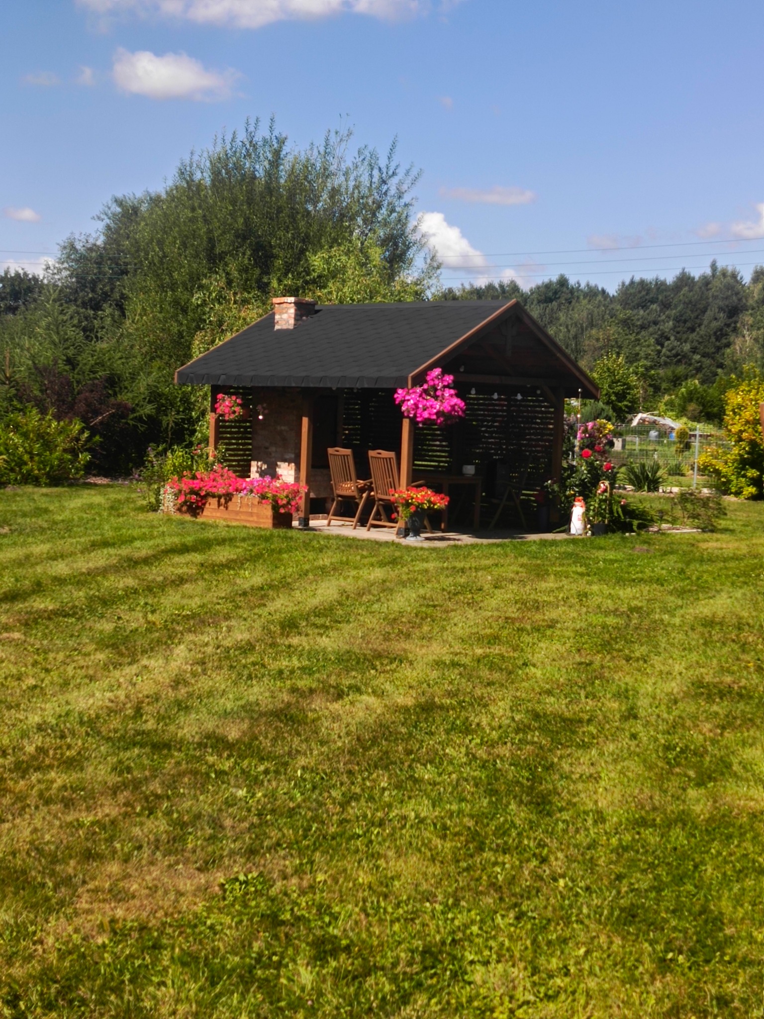 Drewniana altanka z czarnym dachem, zdobiona kwiatami, stoi na trawniku. W tle drzewa i błękitne niebo z chmurami. Sielski krajobraz.