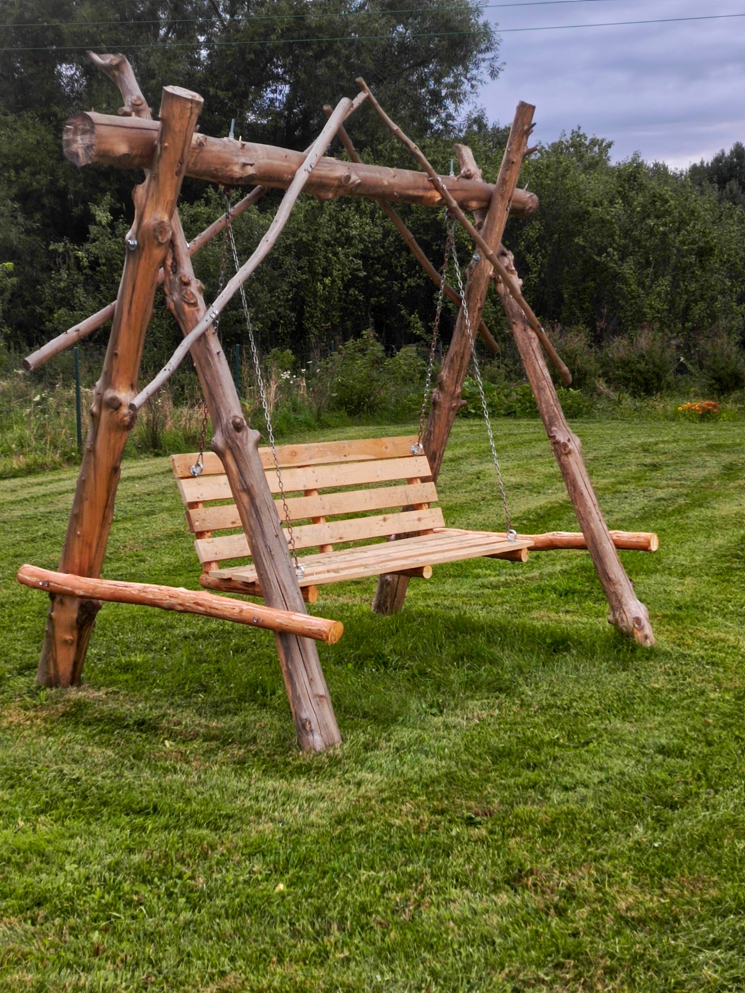 Drewniana huśtawka ogrodowa z surowych bali, zawieszona na łańcuchach, na tle zielonego trawnika i drzew. Naturalny styl, ręczne wykonanie, solidna konstrukcja.