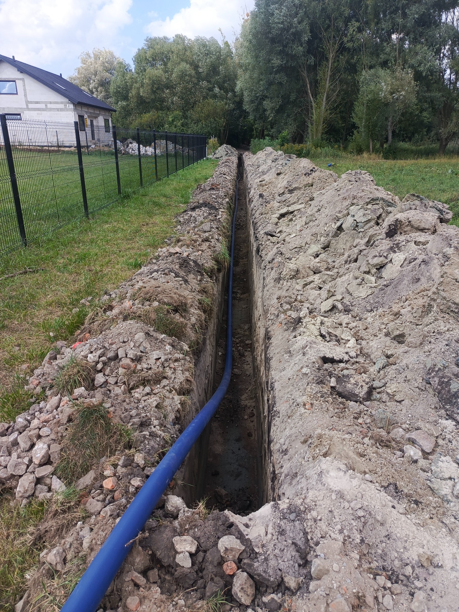 Wąski wykop w ziemi z niebieską rurą wewnątrz, widok z góry. Trawa i ogrodzenie w tle. Wykop świeżo wykonany, z widoczną strukturą ziemi.