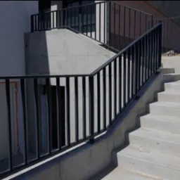 Betonowe schody zewnętrzne z czarną, metalową barierką o pionowych szczeblach. Ujęcie z dołu, widać cień. Minimalistyczny design.