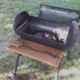 Otwarty grill metalowy z rusztem wypełnionym węglem drzewnym, umieszczony na drewnianym wózku z kółkami, na tle trawnika.