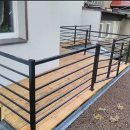 Balkon z drewnianą podłogą i czarną, nowoczesną balustradą z poziomych prętów, widok z góry na zielony dach z drobnymi kamieniami.