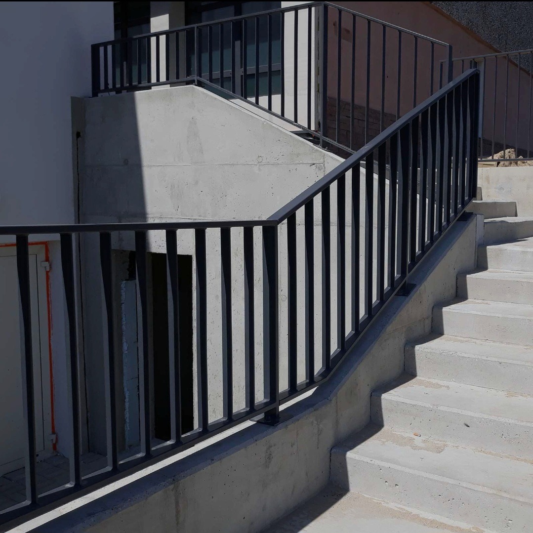 Betonowe schody zewnętrzne z czarną, metalową barierką o pionowych szczeblach. Ujęcie z dołu, widać cień. Minimalistyczny design.