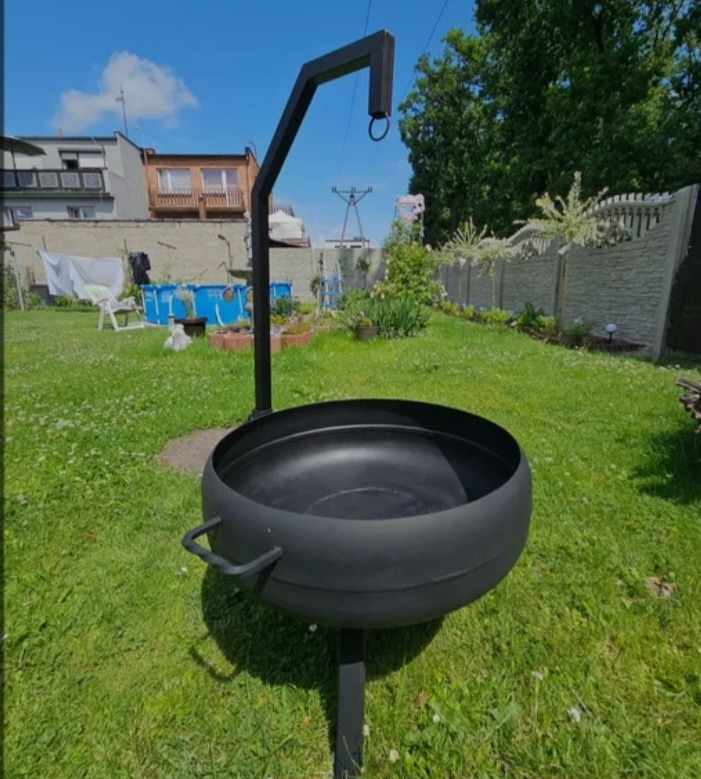 Czarny, metalowy palenisko ogrodowe z uchwytami i ramieniem do zawieszania, ustawione na trawniku. W tle dom, basen i ogrodzenie z betonowych elementów.