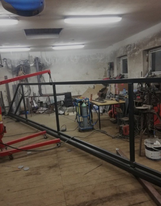 Warsztatowa sceneria z fragmentem konstrukcji metalowej w trakcie tworzenia. Widoczny sprzęt spawalniczy i inne narzędzia ślusarskie w tle.