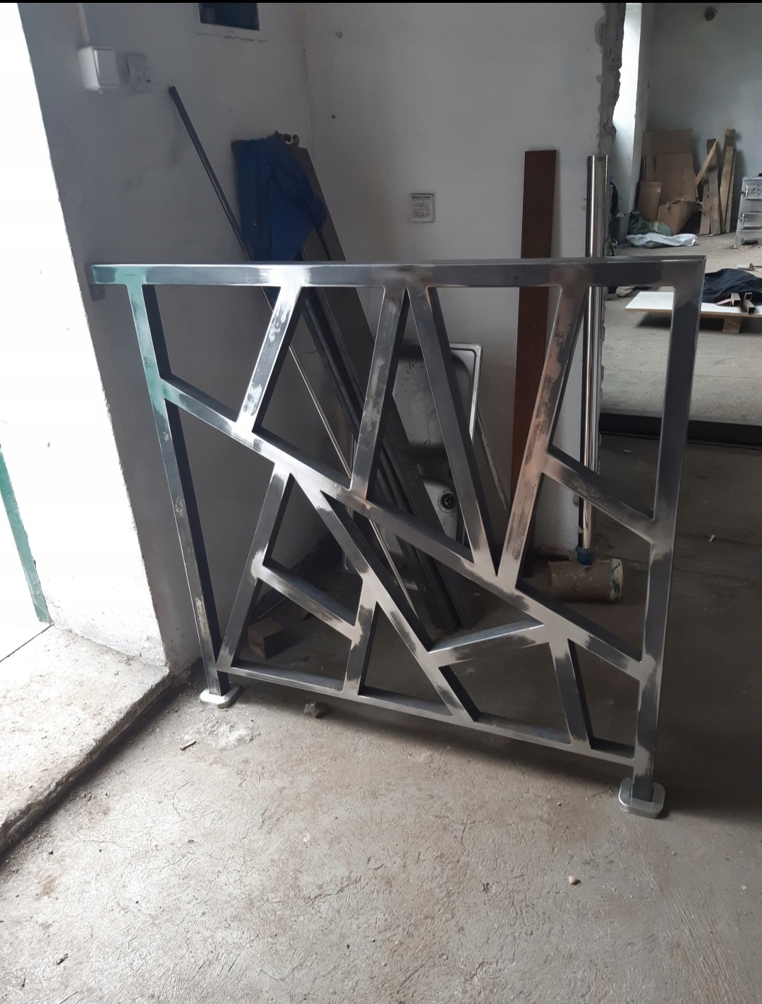 Surowa, metalowa balustrada o geometrycznym wzorze, w trakcie montażu w surowym wnętrzu. Widoczne niedoskonałości spoin i betonowa podłoga.