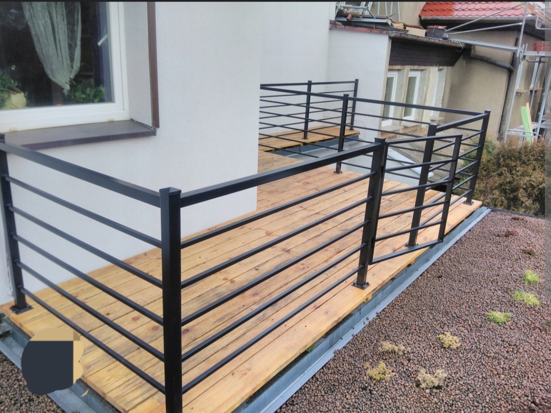 Balkon z drewnianą podłogą i czarną, nowoczesną balustradą z poziomych prętów, widok z góry na zielony dach z drobnymi kamieniami.